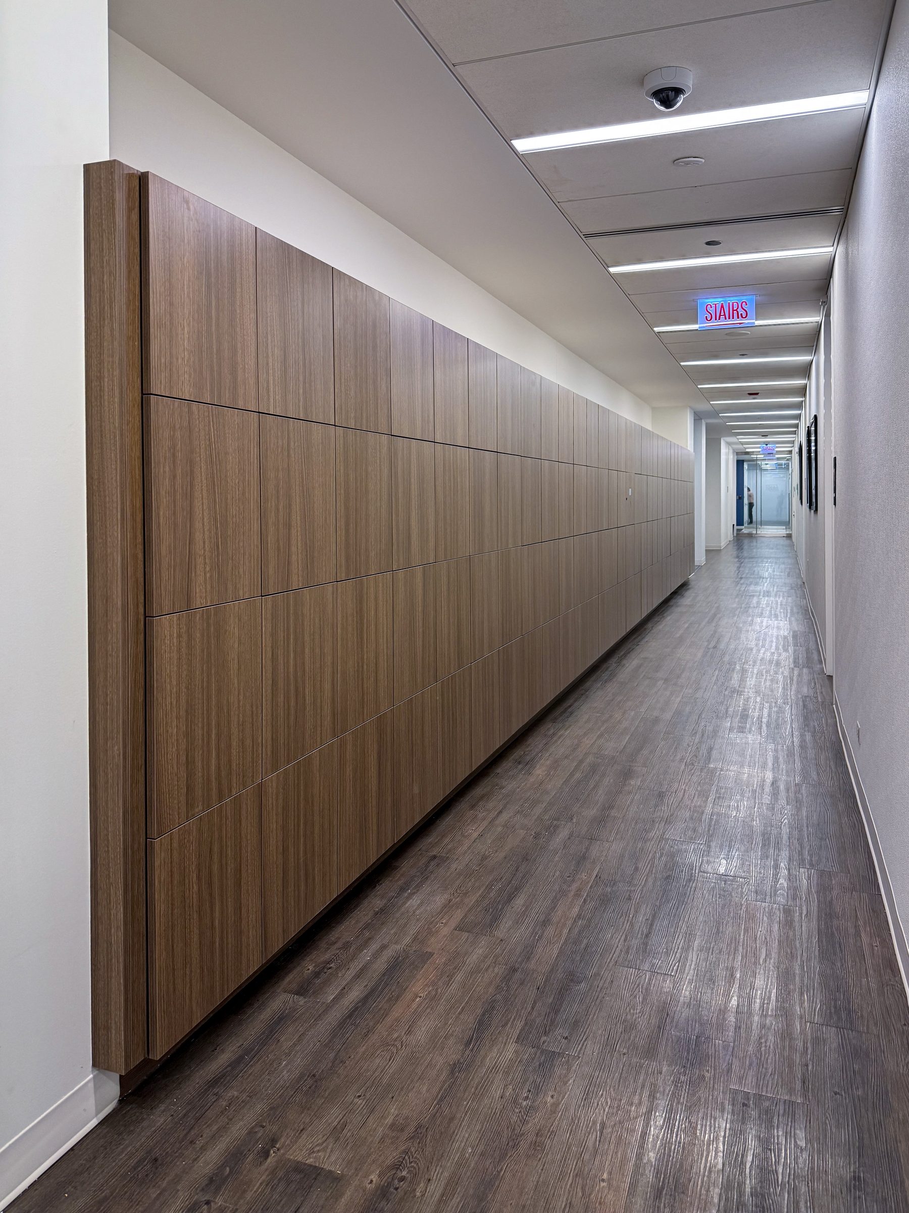 Walnut woodgrain full-height wall lockers running the full length of a corporate office corridor